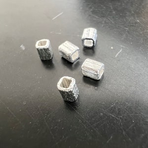 Könnte beinhalten: Fünf silberne quadratische Perlen aus Metall mit strukturierter Oberfläche.