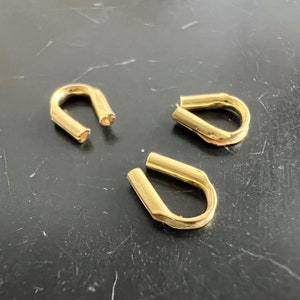 Puede incluir: Tres guardacabos dorados, cada uno con un diseño en forma de U. Los guardacabos están hechos de metal brillante y están dispuestos sobre una superficie oscura. Los extremos abiertos de los guardacabos son visibles.