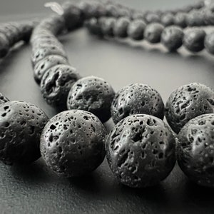 May include: A close-up of a black lava rock bead necklace. The beads are round and have a rough, textured surface. The necklace is made of natural lava rock.
