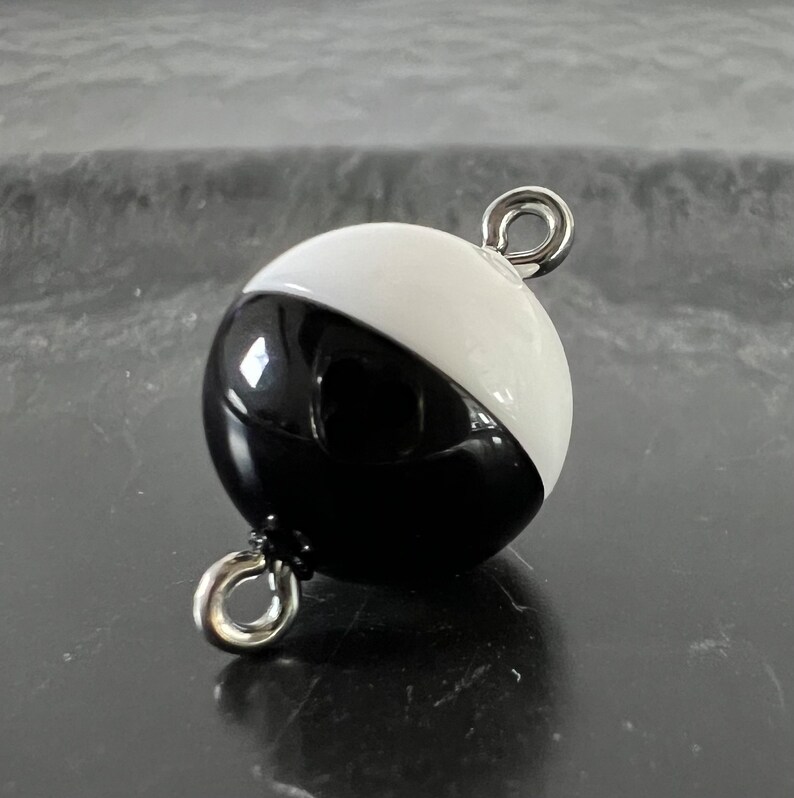 May include: A black and white round ornament with a silver metal loop on each side. The ornament is half black and half white.