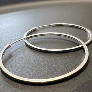 Pode incluir: Um par de brincos de argola prateados com um design de fio fino e circular. Os brincos apresentam uma construção simples de banda metálica contínua com um acabamento liso e polido que reflete a luz, criando uma aparência limpa e discreta.
