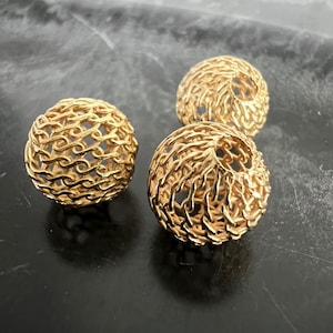 Könnte beinhalten: Drei goldfarbene Metallkugeln mit einem Kettenglied-Design. Die Kugeln sind in einem dreieckigen Muster auf einer dunkelgrauen Oberfläche angeordnet.