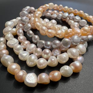 Peut inclure: Un ensemble de trois bracelets en perles de différentes nuances de rose, blanc et gris. Les perles sont grandes et de forme irrégulière, ce qui donne aux bracelets un aspect unique et élégant.