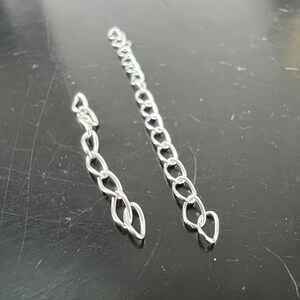 May include: Two silver chains, one longer than the other, are shown against a dark background. The chains are made of small, linked oval shapes.