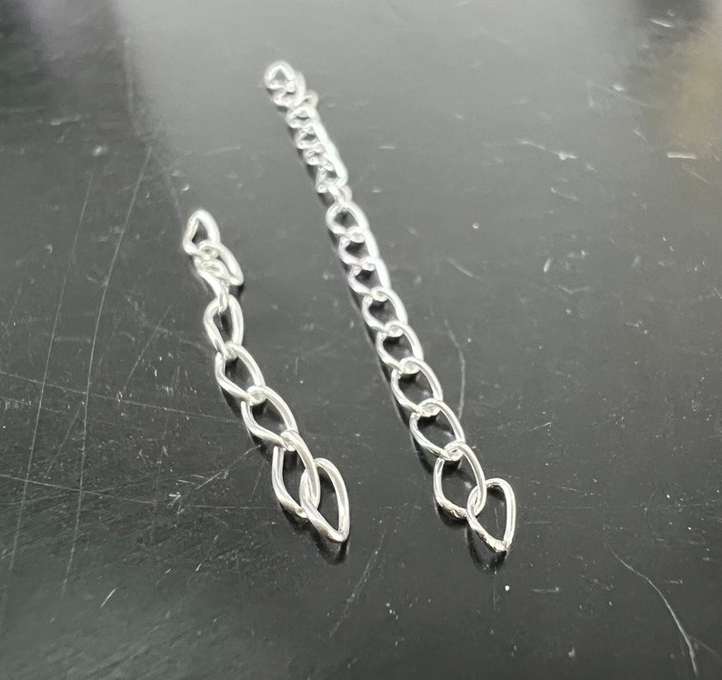 May include: Two silver chains, one longer than the other, are shown against a dark background. The chains are made of small, linked oval shapes.