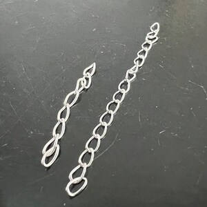 May include: Two silver chains, one longer than the other, are laid out on a dark gray surface.