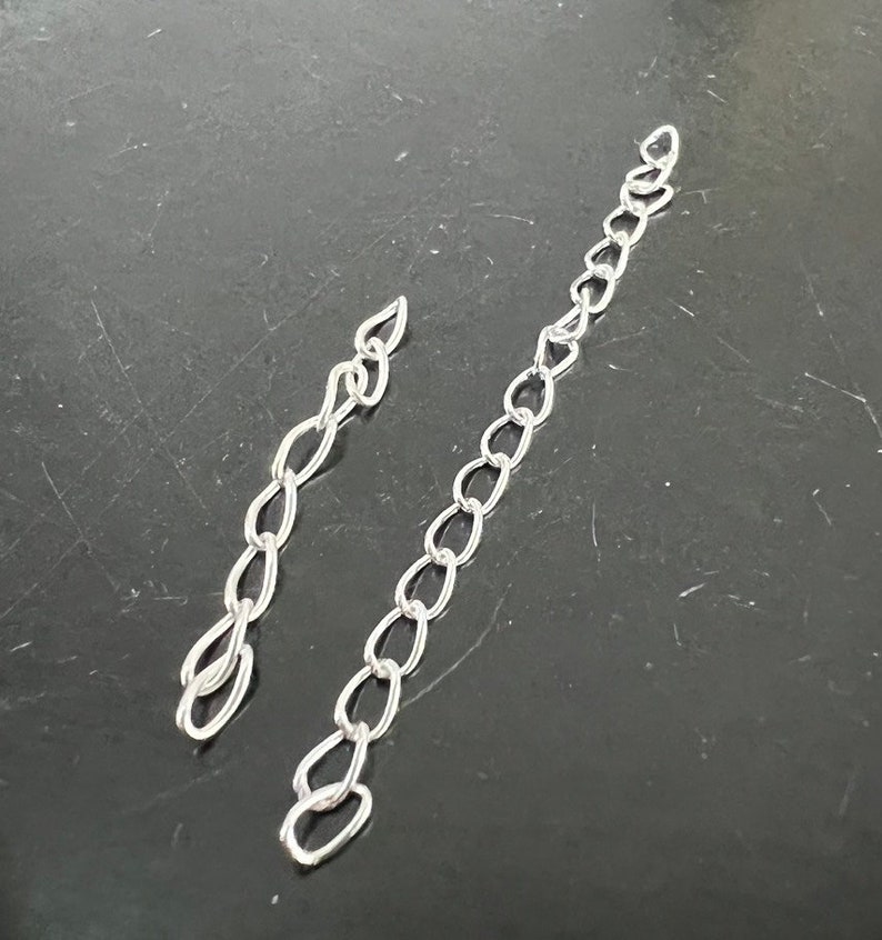 May include: Two silver chains, one longer than the other, are laid out on a dark gray surface.
