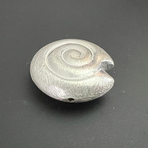 Może przedstawiać: Srebrny wisiorek w kształcie muszli z spiralnym wzorem. Wisiorek ma otwór do przymocowania łańcuszka.