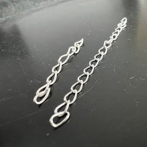 May include: Two silver chains, one longer than the other, are laid out on a black surface. The chains are made of small, oval-shaped links.