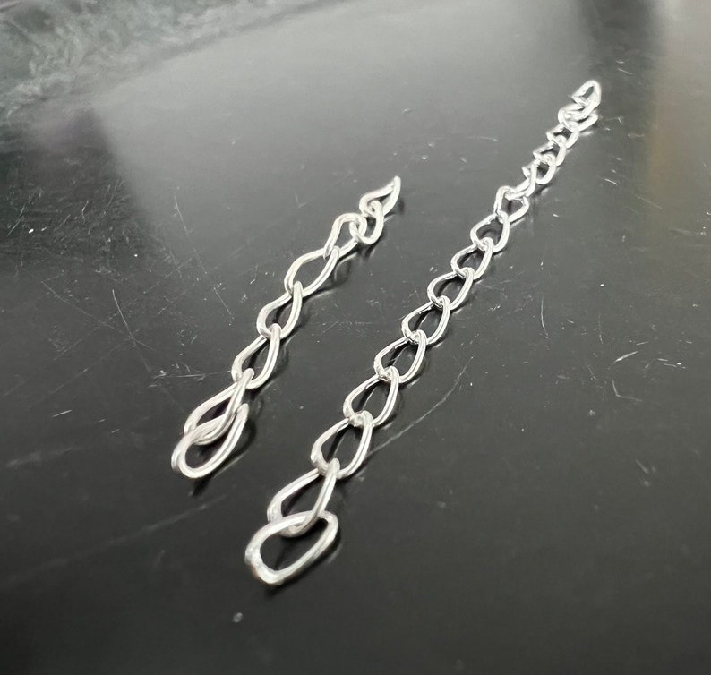 May include: Two silver chains, one longer than the other, are laid out on a black surface. The chains are made of small, oval-shaped links.