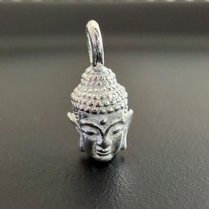 Könnte beinhalten: Ein silberfarbener Metallanhänger in Form eines Buddha-Kopfes. Der Anhänger zeigt eine strukturierte, abgerundete Oberseite, die das Haar darstellt, verlängerte Ohrläppchen, geschlossene Augen und einen kleinen Punkt auf der Stirn. Oben befindet sich eine kleine Metallöse.