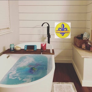 May include: A white bathtub filled with blue water and bubbles. A wooden bath tray with a video game console, a glass of red liquid, and a can of beer is placed on the edge of the tub. The bathroom has white shiplap walls and a wooden floor.