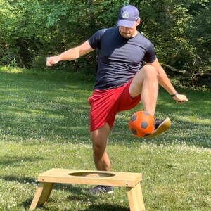 May include: A person wearing a blue baseball cap and a red and black shirt is kicking a soccer ball into a wooden ring toss game.