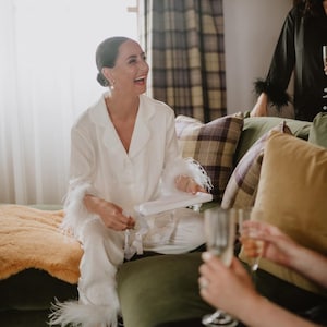 May include: A woman wearing a white satin pyjama set with feather trim sits on a green sofa and laughs. She is holding a white box and there are two glasses of champagne on the table in front of her.