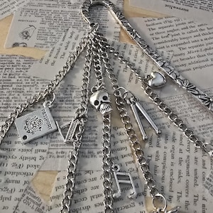 May include: A silver bookmark with a chain attached to various charms, including a skull, a heart, a passport, a music note, and a drum. The bookmark is decorated with a floral design.