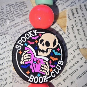 Puede incluir: Un collar de cuentas con un colgante negro circular con un cráneo leyendo un libro y las palabras "Spooky Book Club". El collar tiene cuentas de color rosa brillante y verde menta.