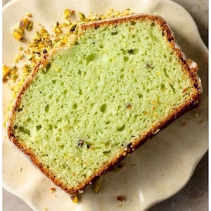 May include: A slice of pistachio cake with a light green hue, topped with a white glaze and chopped pistachios. The cake sits on a cream-colored plate, with scattered pistachio pieces around it. The cake has a moist texture.
