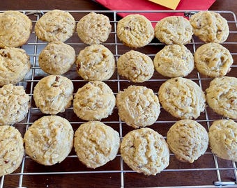 May include: A rectangular metal wire cooling rack holds twenty-four round, golden-brown baked cookies with visible textured edges and small dark inclusions, arranged in a grid of four rows and six columns on a dark surface.