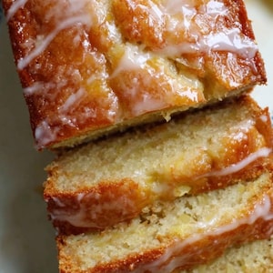 May include: A sliced loaf of golden-brown cake with a white, glossy glaze drizzled over the top. The cake has a moist appearance, with visible textures and a light, airy crumb. The glaze adds a sweet, appealing contrast to the cake.