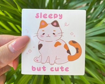 May include: A sticker featuring a cartoon calico cat with orange, black, and white markings. The cat is sitting with its eyes closed and a sleepy expression. The text "sleepy but cute" is written below the cat.