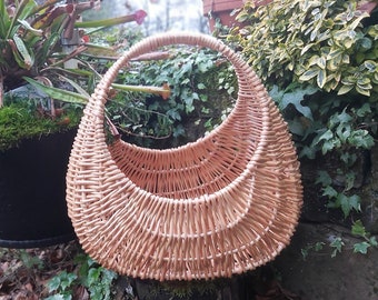 May include: A natural woven wicker basket with a rounded, crescent shape and a single handle. The basket is light brown and sits on a tree stump.