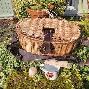 May include: A woven wicker picnic basket with a brown leather strap and clasp. A white enamel mug with a red design, a wooden brush, and a wooden knife rest on a mossy surface.