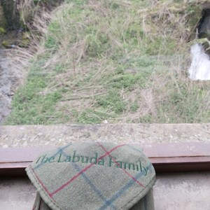 May include: A green tweed case with a flap, displaying a plaid pattern with red and blue lines. The text "The Labuda Family" is embroidered in green on the flap. The case is set against a natural outdoor backdrop.