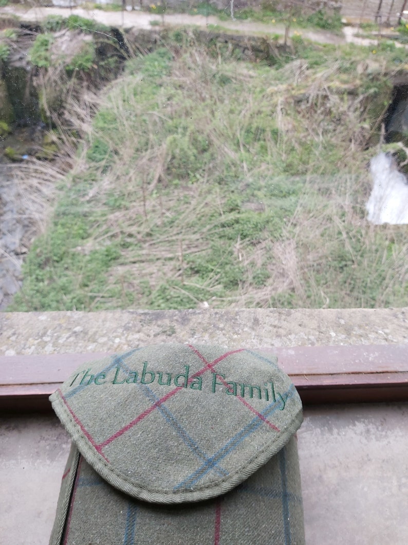 May include: A green tweed case with a flap, displaying a plaid pattern with red and blue lines. The text "The Labuda Family" is embroidered in green on the flap. The case is set against a natural outdoor backdrop.