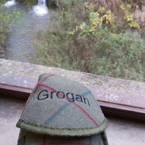 May include: A green and red plaid flask cover with the name "Grogan" embroidered in black. The cover has a green trim and a strap. The background shows a small waterfall and greenery.