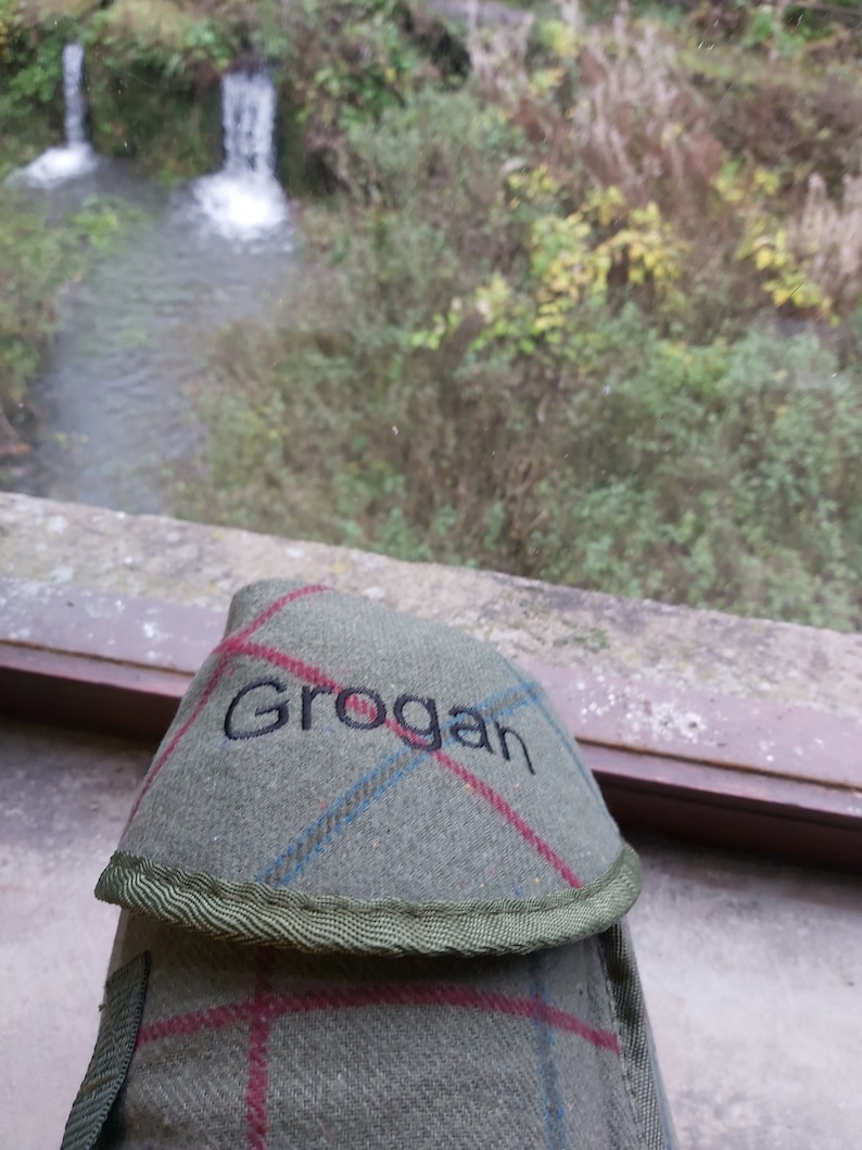 May include: A green and red plaid flask cover with the name "Grogan" embroidered in black. The cover has a green trim and a strap. The background shows a small waterfall and greenery.