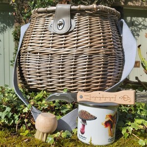 May include: A wicker basket with a leather handle and a gray strap, a mushroom identification guide, a mushroom brush, and a small knife with a ruler on the handle.