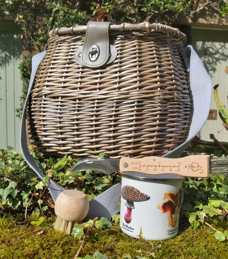 May include: A wicker basket with a leather handle and a gray strap, a mushroom identification guide, a mushroom brush, and a small knife with a ruler on the handle.