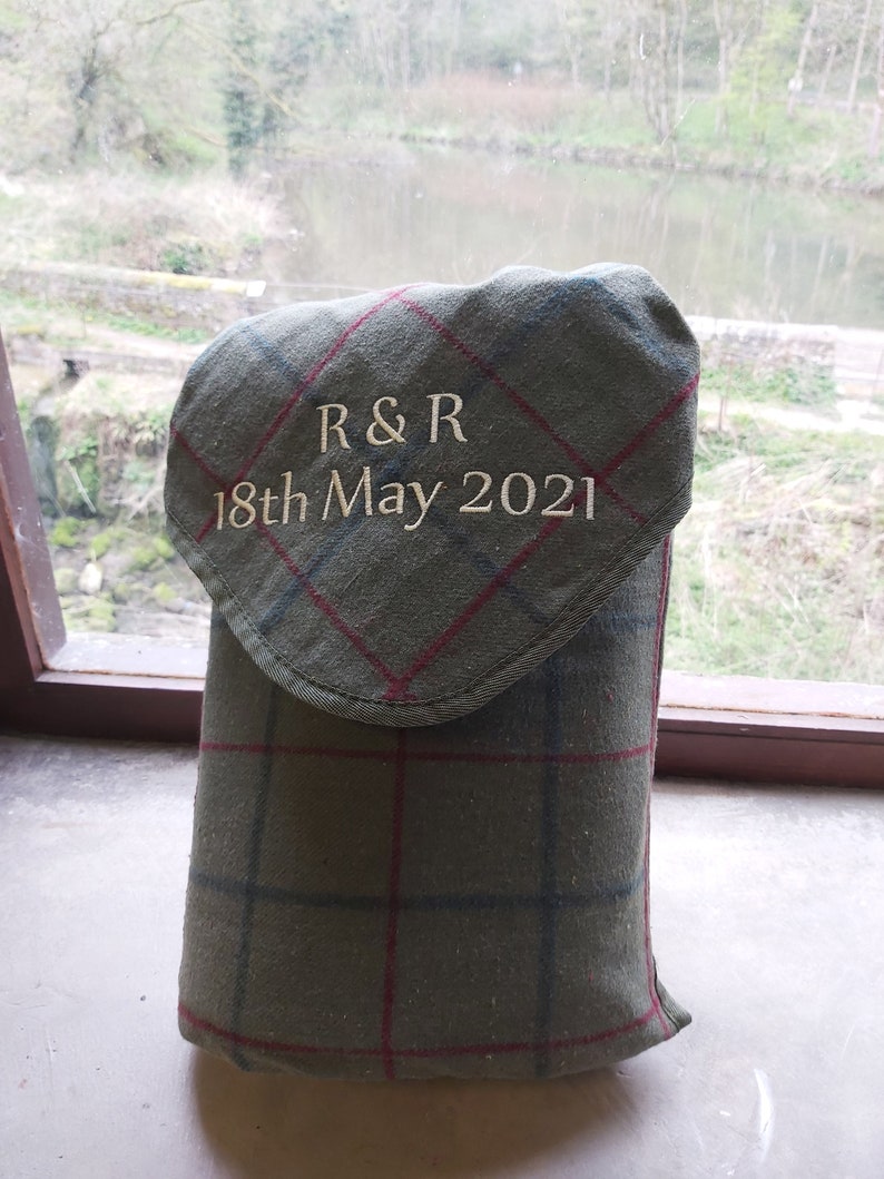 May include: A green plaid fabric item with a flap, embroidered with "R & R" and "18th May 2021". The item is likely a case or cover, possibly for a bottle or other small object. The fabric has red and blue lines.