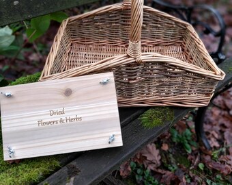 May include: A wicker basket with a wooden handle sits on a wooden bench. A wooden sign with the text "Dried Flowers & Herbs" is in front of the basket.