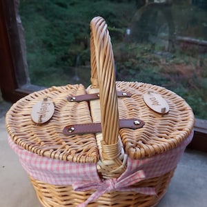 May include: A wicker picnic basket with a pink and white checkered lining and a brown leather handle. The basket has two lids with wooden name tags that say "Evelyn" and "Ava".