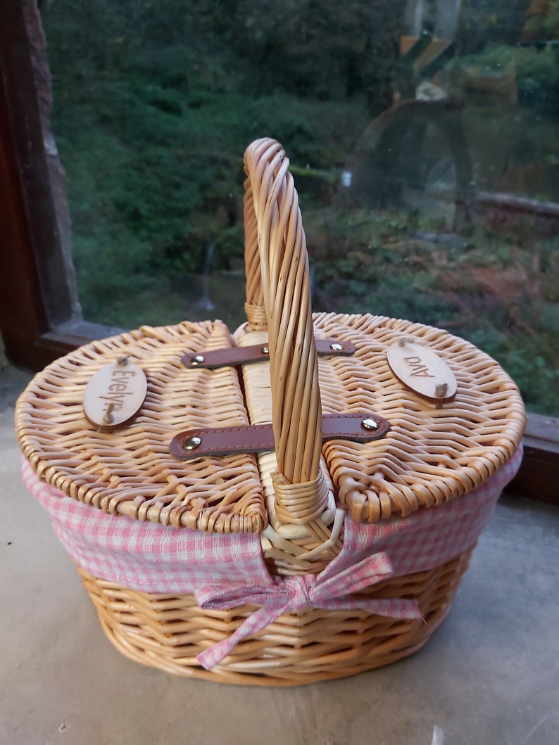 May include: A wicker picnic basket with a pink and white checkered lining and a brown leather handle. The basket has two lids with wooden name tags that say "Evelyn" and "Ava".