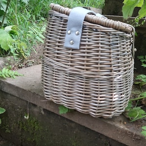 May include: A gray wicker bicycle basket with a gray fabric strap and silver metal accents. The basket is sitting on a stone step.