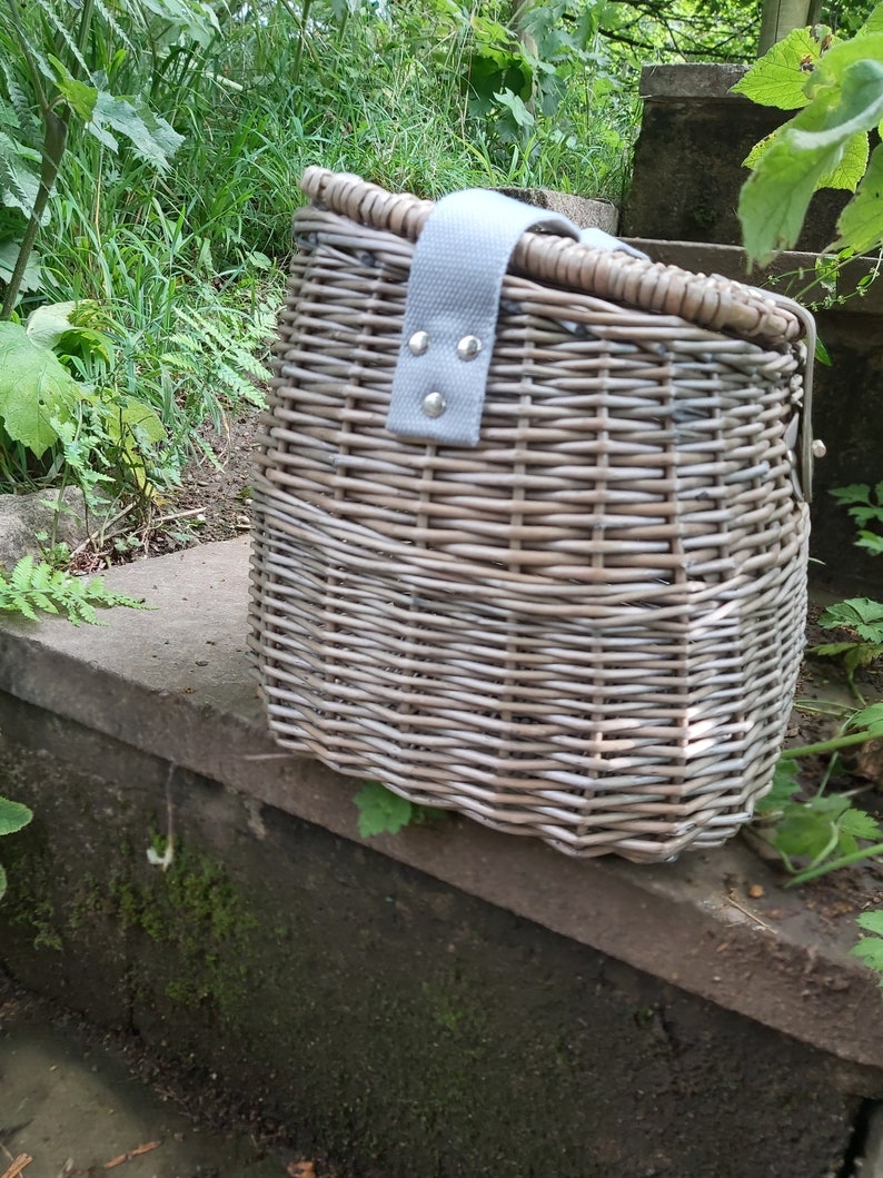 May include: A gray wicker bicycle basket with a gray fabric strap and silver metal accents. The basket is sitting on a stone step.
