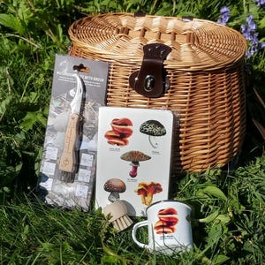 May include: A woven wicker picnic basket with a brown leather closure, a mushroom knife with a brush in its original packaging, a white notebook featuring various mushroom illustrations, and a white enamel mug with a mushroom graphic print.