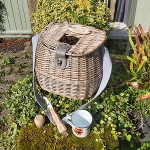 May include: A wicker fishing basket with a leather strap and a wooden sign that reads "The best days are spent between you and the water". The basket is sitting on a bed of green moss with a white enamel mug and a wooden brush.