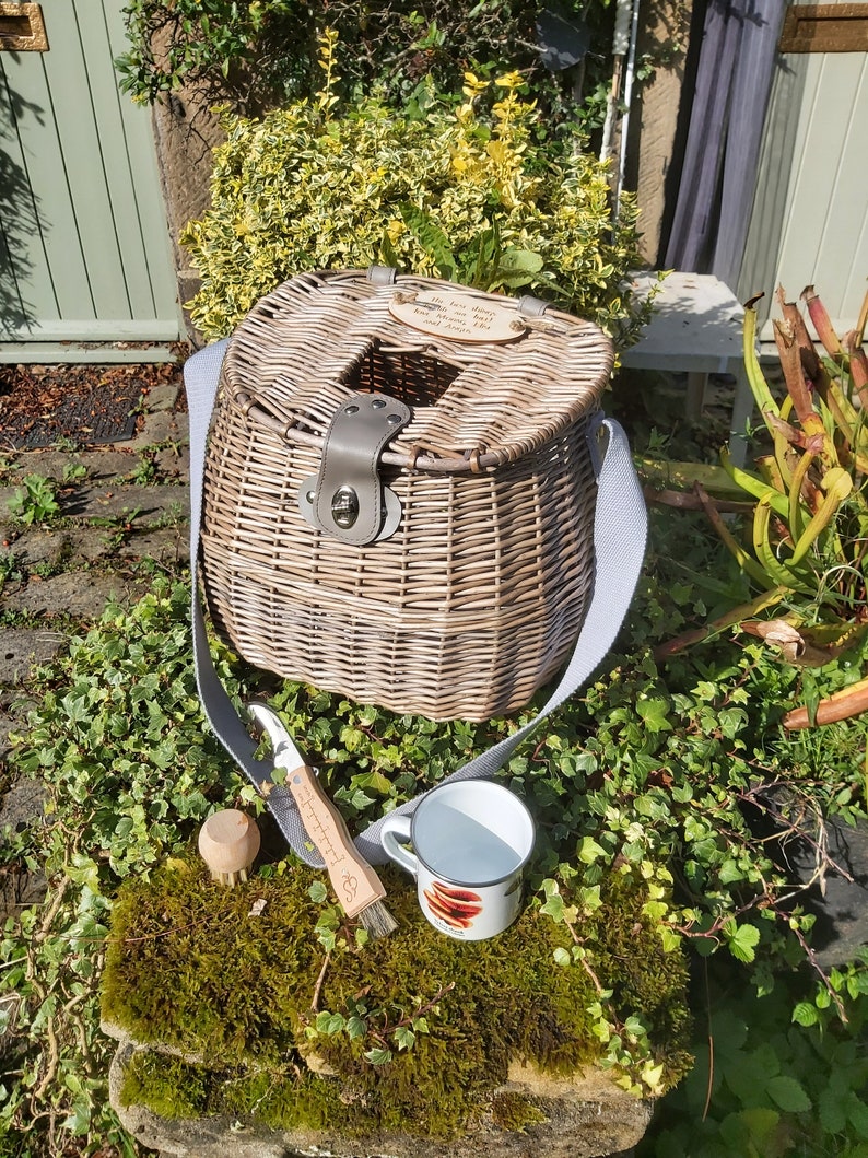 May include: A wicker fishing basket with a leather strap and a wooden sign that reads "The best days are spent between you and the water". The basket is sitting on a bed of green moss with a white enamel mug and a wooden brush.