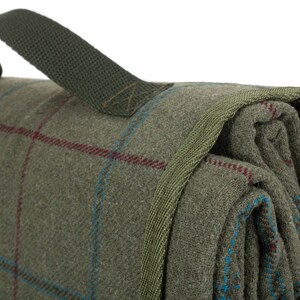May include: Folded green plaid blanket with a green carry handle. The blanket has a woven texture with red and blue accent lines. The handle is made of a durable, woven material.