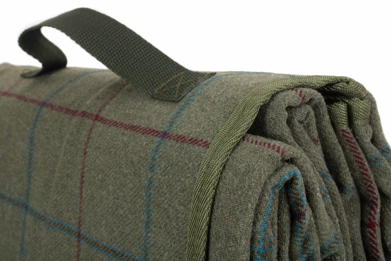 May include: Folded green plaid blanket with a green carry handle. The blanket has a woven texture with red and blue accent lines. The handle is made of a durable, woven material.