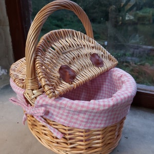 May include: A wicker picnic basket with a pink and white checkered lining. The basket has a hinged lid and a brown leather handle.