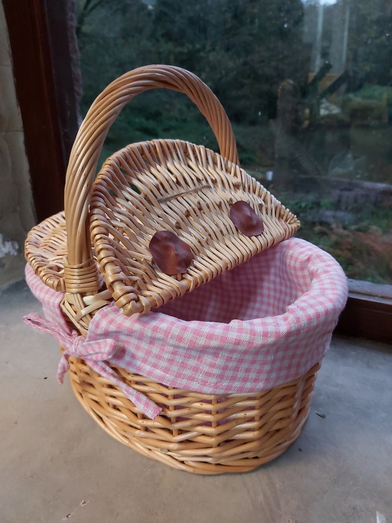May include: A wicker picnic basket with a pink and white checkered lining. The basket has a hinged lid and a brown leather handle.