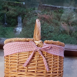 May include: A wicker picnic basket with a pink and white gingham fabric lining and a bow. The basket has a handle and is made of natural woven wicker.
