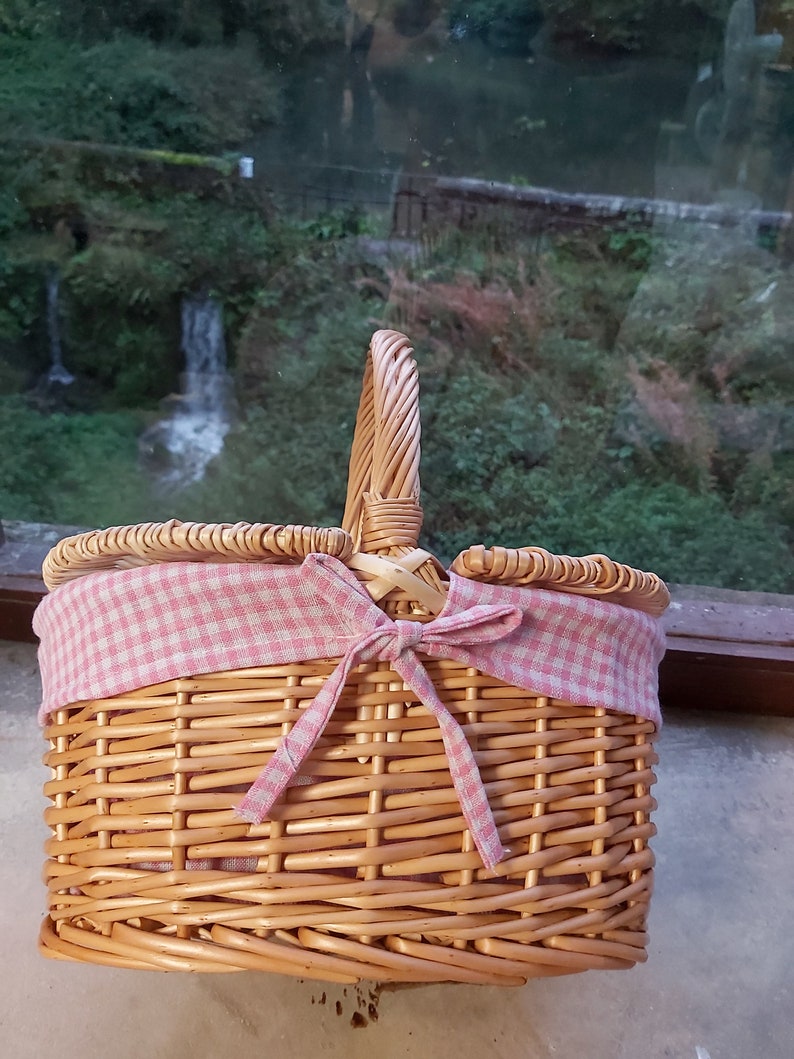 May include: A wicker picnic basket with a pink and white gingham fabric lining and a bow. The basket has a handle and is made of natural woven wicker.