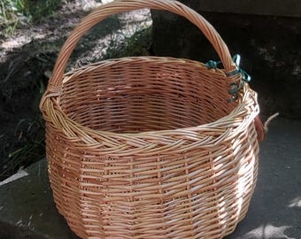 May include: A round, light brown woven wicker basket featuring a single arched handle attached to the sides. The basket has a sturdy, circular base and a braided rim design, constructed from natural willow reeds with a visible, textured weave pattern.