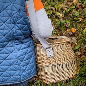 May include: A wicker basket with a gray strap, worn over the shoulder. The basket is brown and has a woven pattern. The strap has a silver buckle.