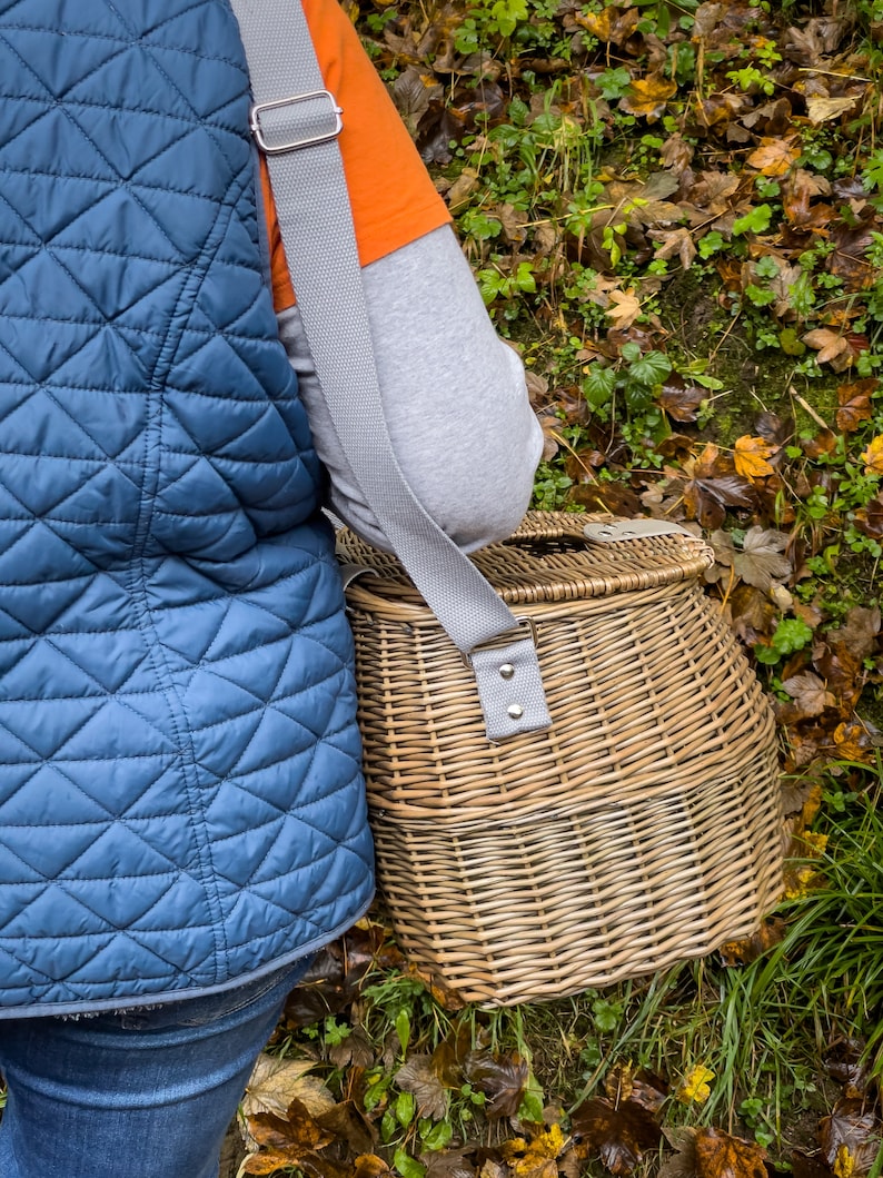 May include: A wicker basket with a gray strap, worn over the shoulder. The basket is brown and has a woven pattern. The strap has a silver buckle.